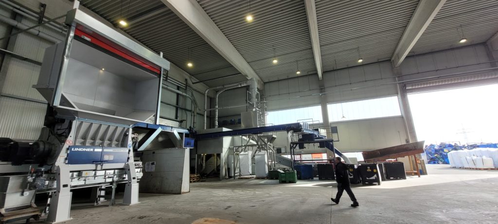 Industrial processing warehouse with a large Lindner machine, conveyors, and a worker walking across the concrete floor.