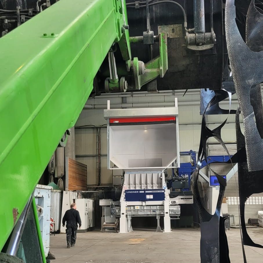 Large green hydraulic/crane arm dominates an industrial workshop, with a worker walking toward a blue processing unit in the background.