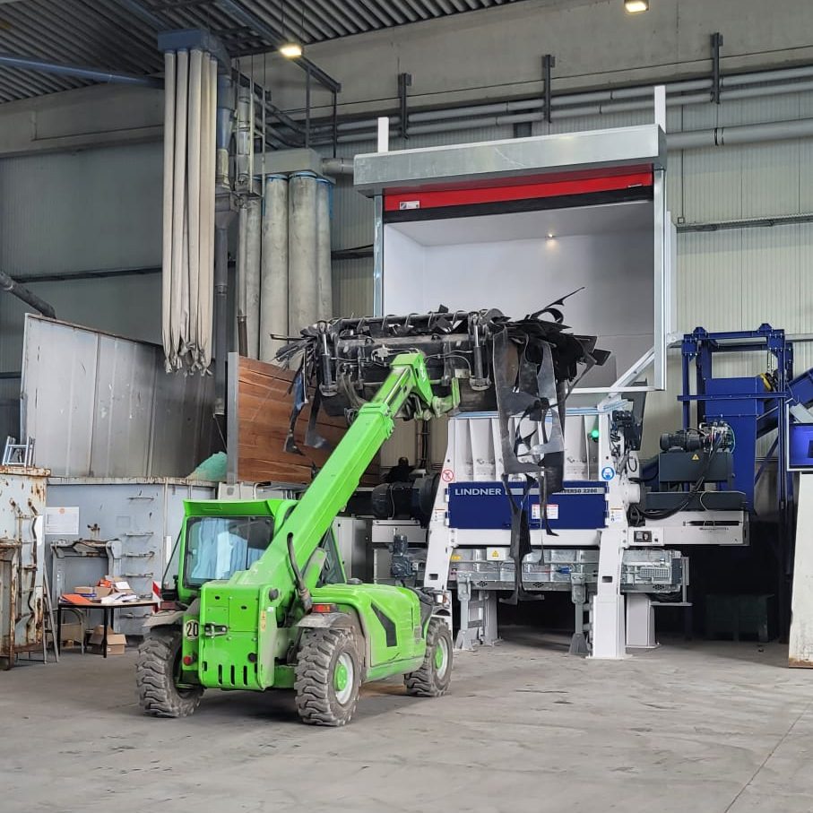 Green telescopic forklift moving scrap into a large industrial machine inside a workshop; metal beams and skylights above.