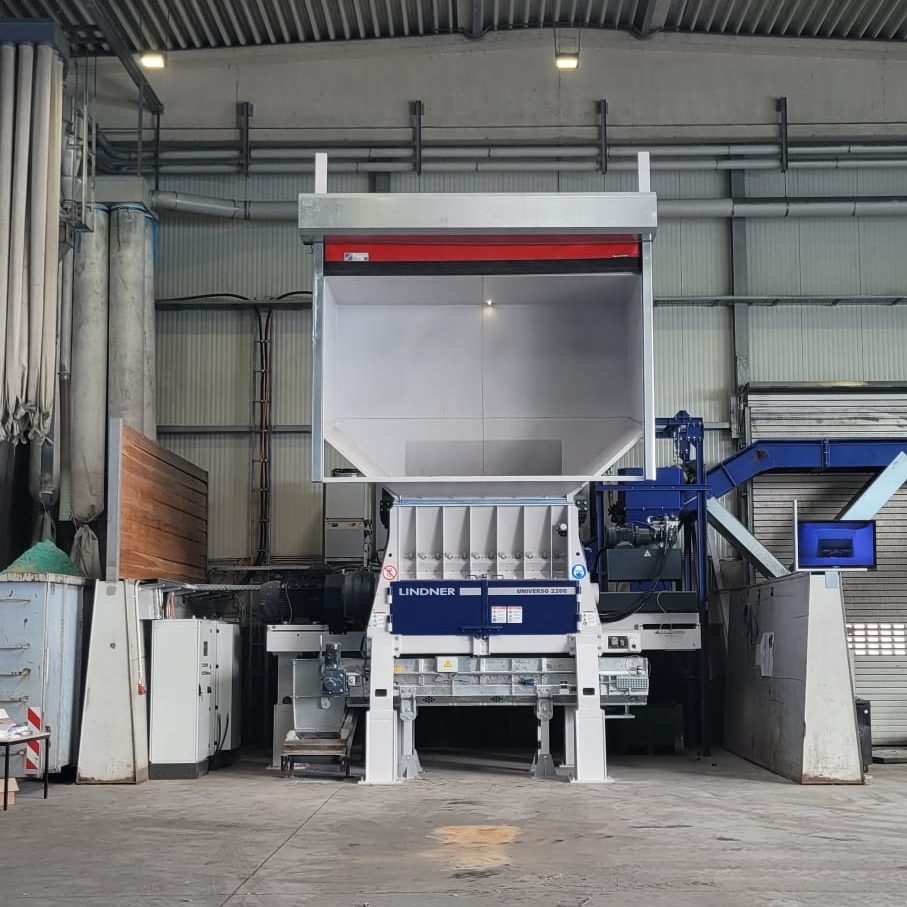 Large industrial machine in a factory, white and blue frame with a tall hopper at the center and overhead skylight above.