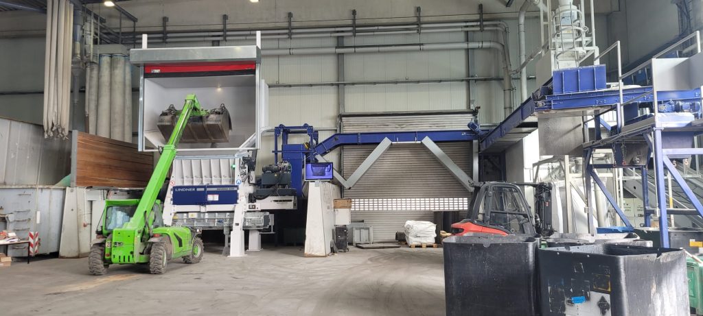 Industrial warehouse with green telescopic forklift lifting a large metal hopper into a white machine enclosure