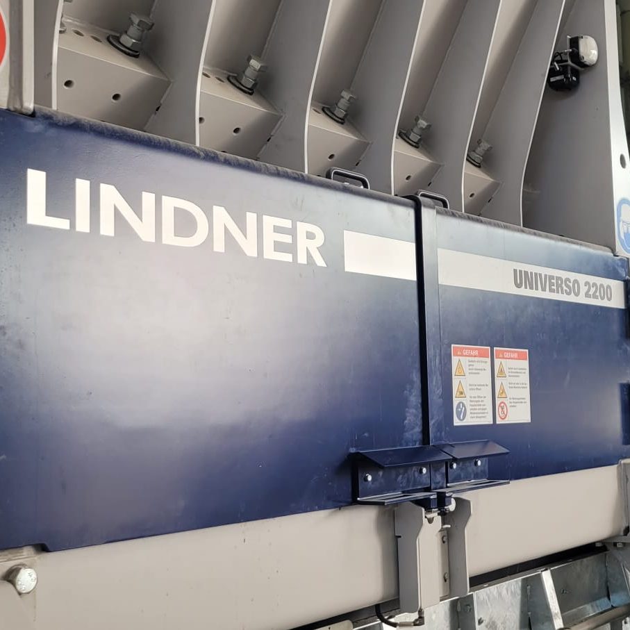 Large blue Lindner machine labeled Universo 2200 in a factory, with warning signs and protective hinges visible on the door.
